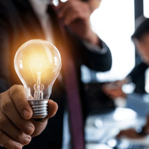 A man wearing a suit holds a glowing lightbulb up to the camera, behind him are two more men working together on a project.