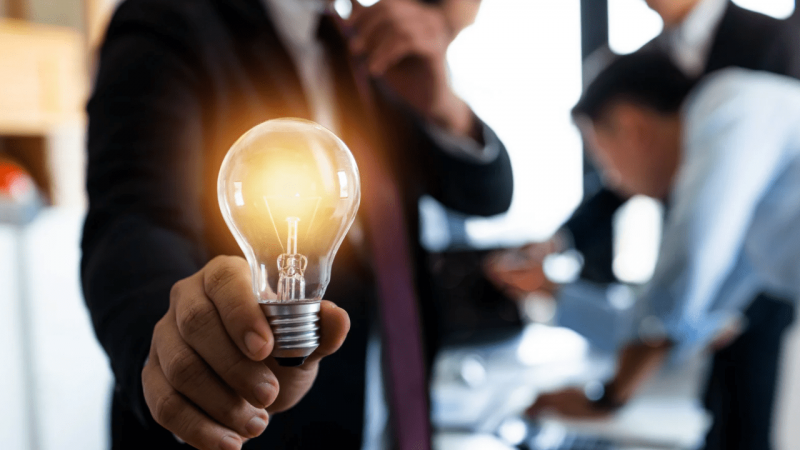 A man wearing a suit holds a glowing lightbulb up to the camera, behind him are two more men working together on a project.